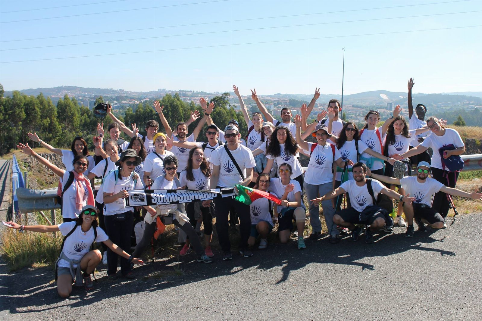 Large group of young people in white T-shirts with a scallop shell, arms raised, holding a Portuguese flag and a “Somos únicos” scarf