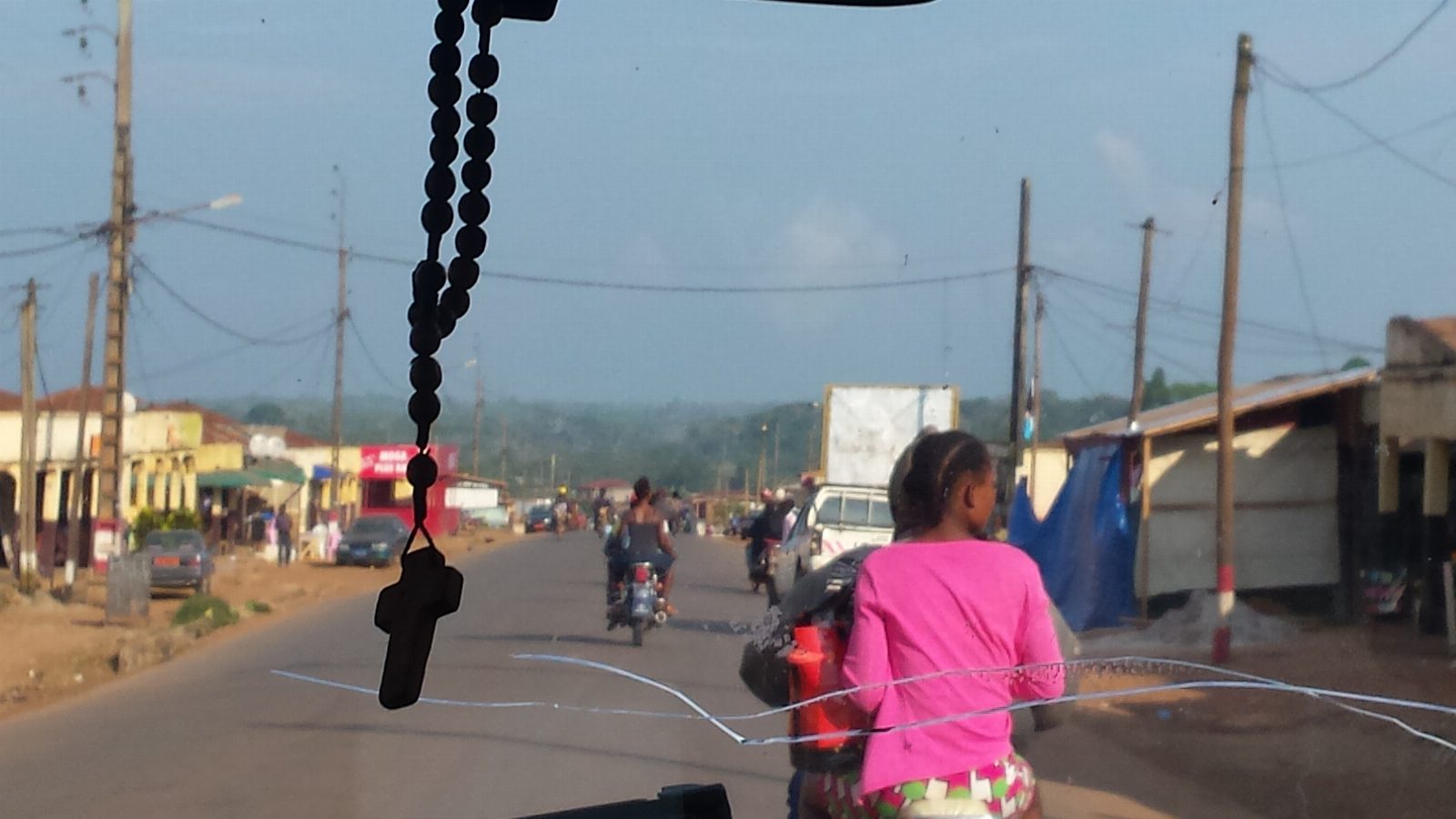 Street in Yaoundé, Cameroon, seen through a windshield with a rosary hanging from the mirror, with motorbikes and a girl in pink