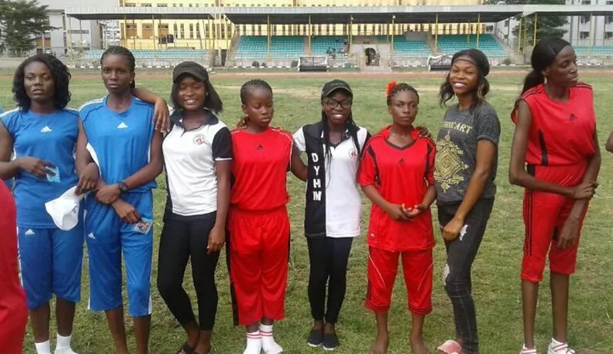 Young women in sports kits, one in a DYM shirt, stand in a row on a sports field in Nigeria